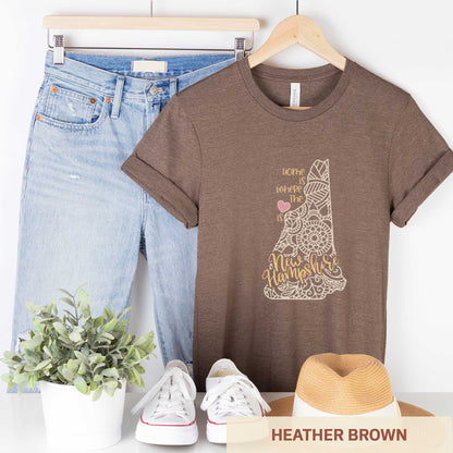 A hanging heather brown Bella Canvas t-shirt featuring a mandala in the shape of New Hampshire with the words home is where the heart is.