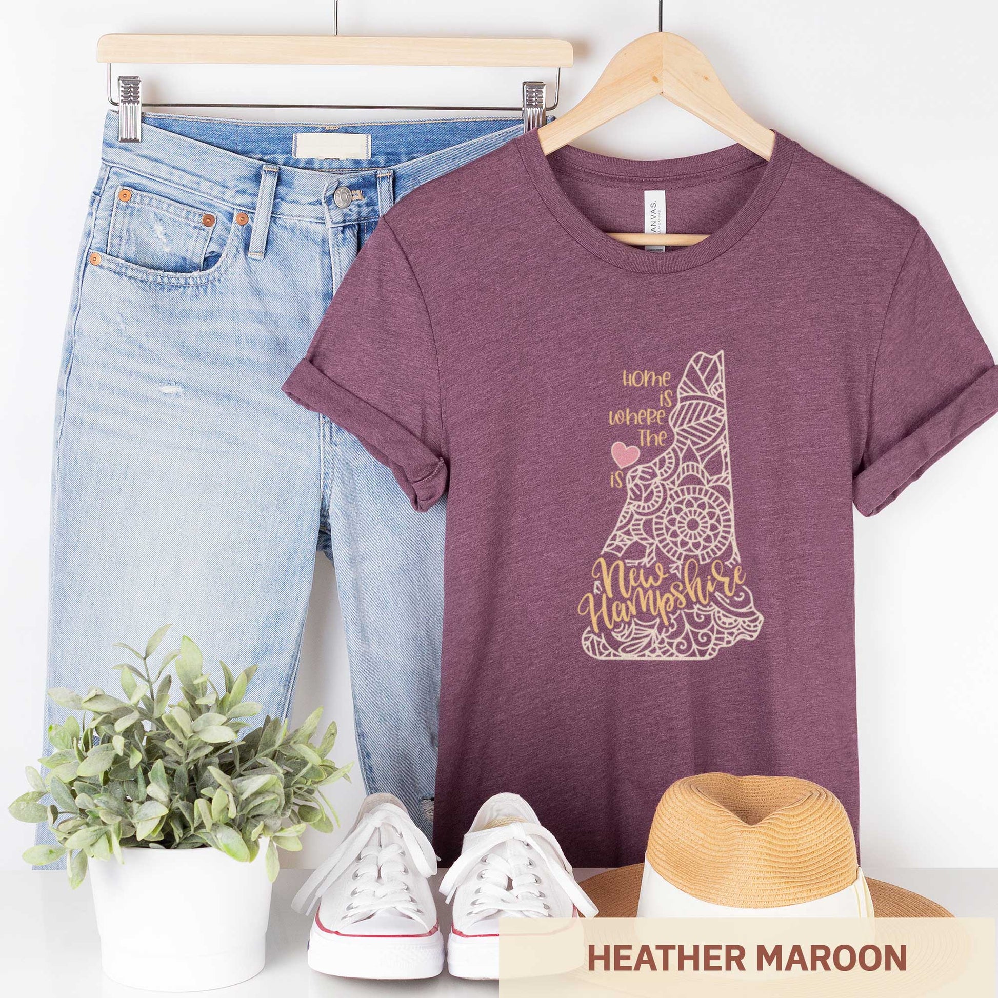 A hanging heather maroon Bella Canvas t-shirt featuring a mandala in the shape of New Hampshire with the words home is where the heart is.