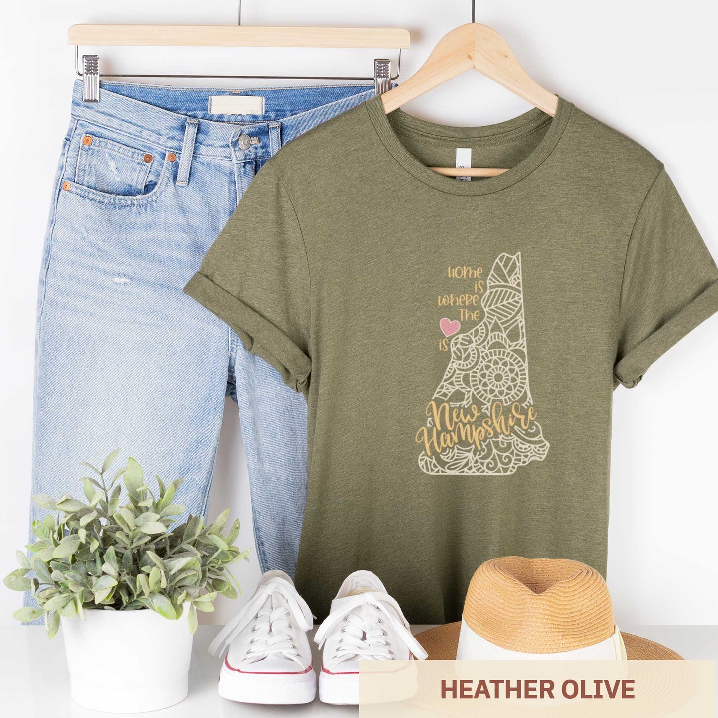 A hanging heather olive Bella Canvas t-shirt featuring a mandala in the shape of New Hampshire with the words home is where the heart is.