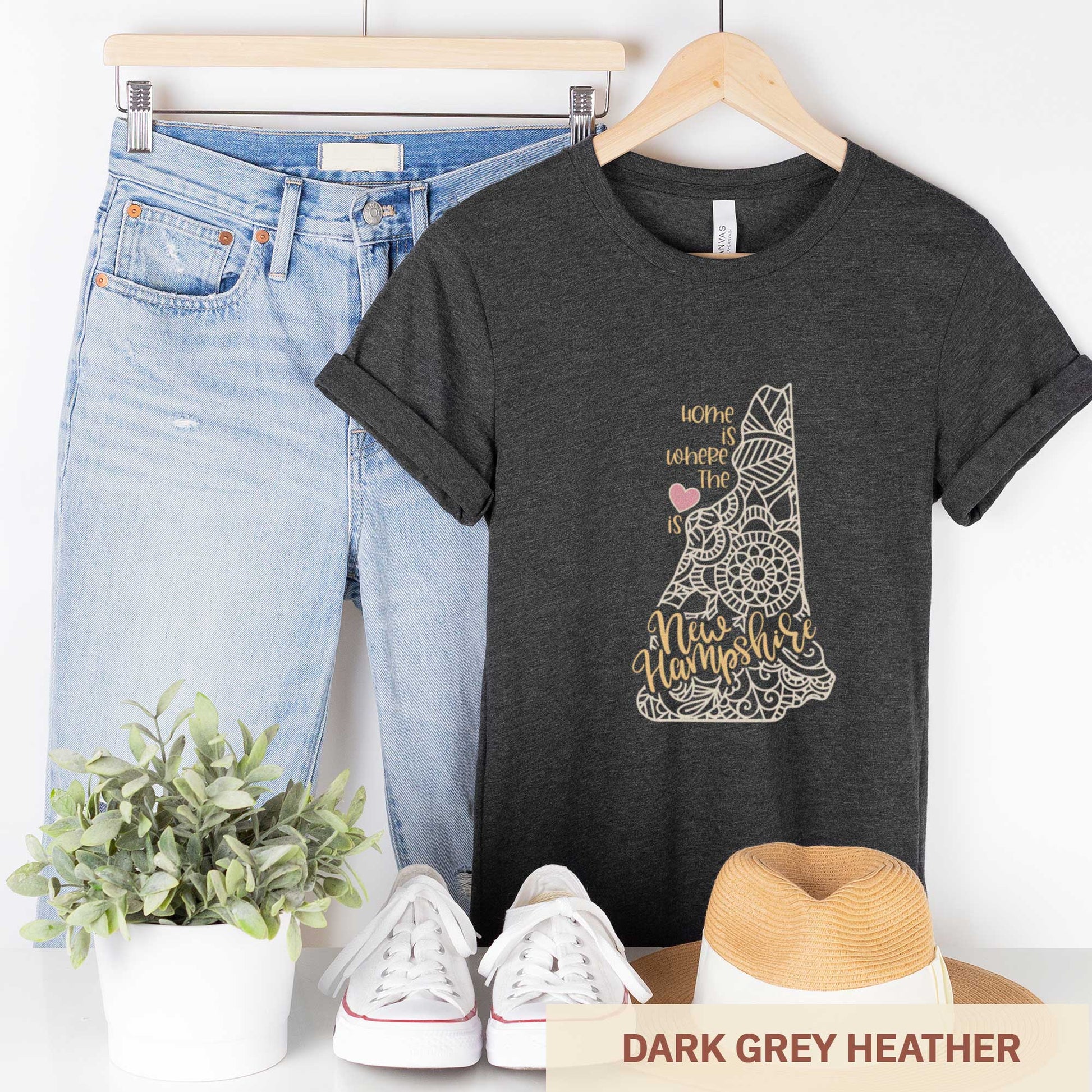 A hanging dark grey heather Bella Canvas t-shirt featuring a mandala in the shape of New Hampshire with the words home is where the heart is.