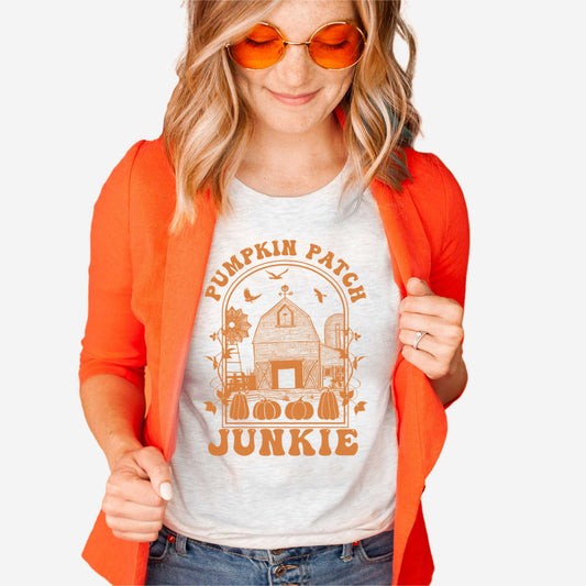 A woman wearing an ash Bella Canvas t-shirt featuring a farm with pumpkins that says pumpkin patch junkie.