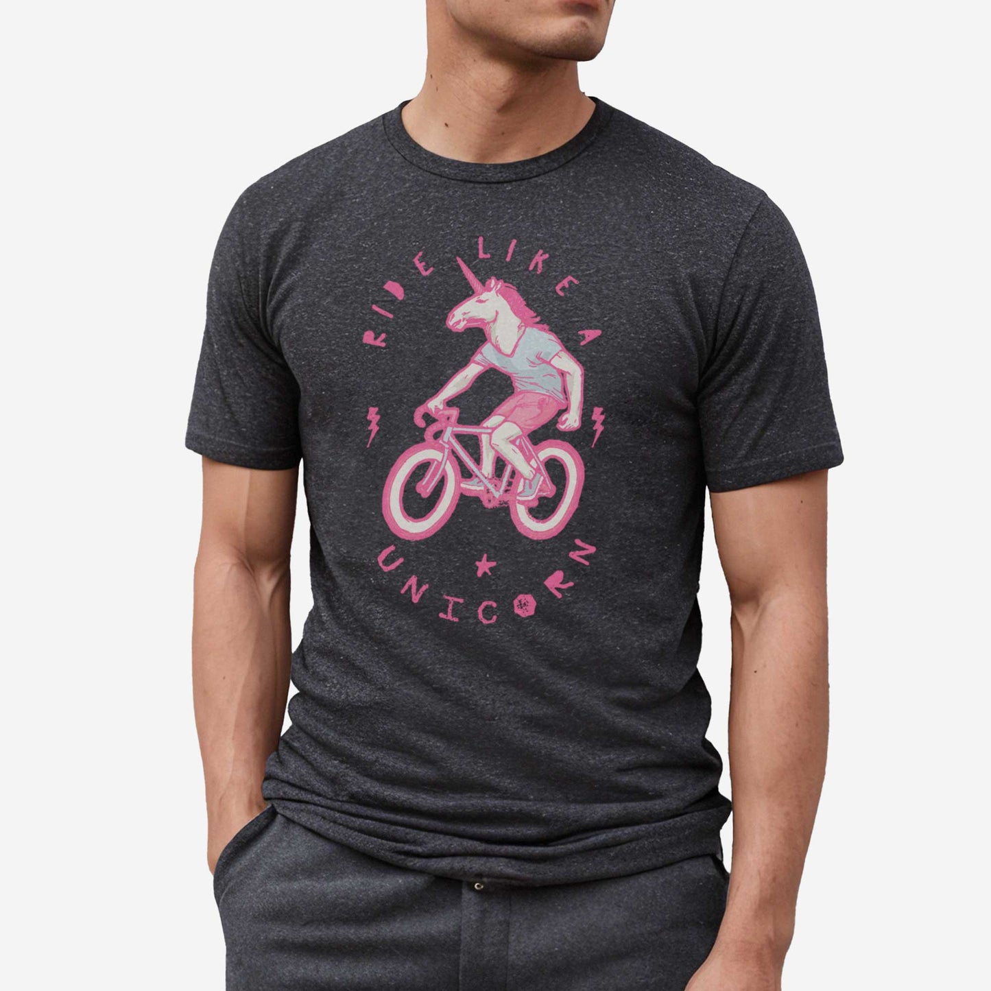A man wearing a dark grey heather Bella Canvas t-shirt featuring a neon pink unicorn riding a bike and the words ride like a unicorn.