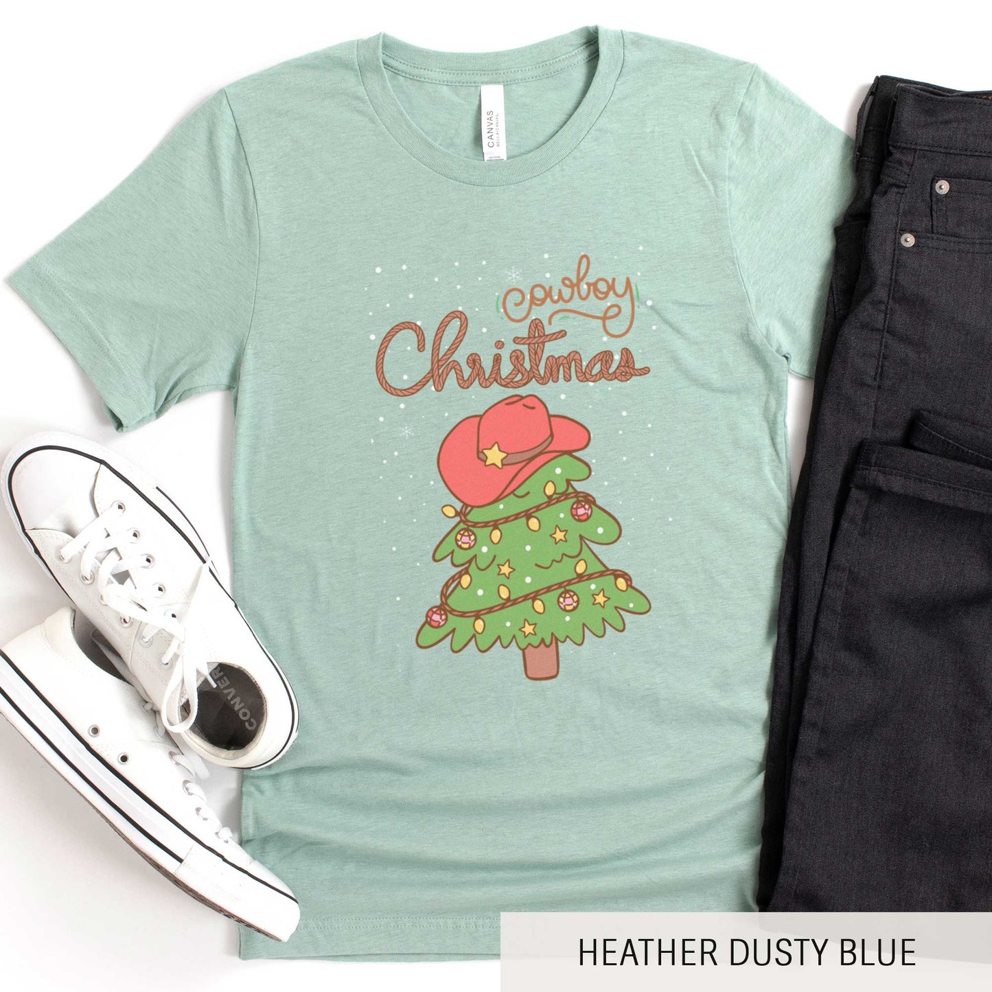 A heather dusty blue Bella Canvas t-shirt featuring a cartoon christmas tree with the words cowboy christmas above it and snow falling in the background.