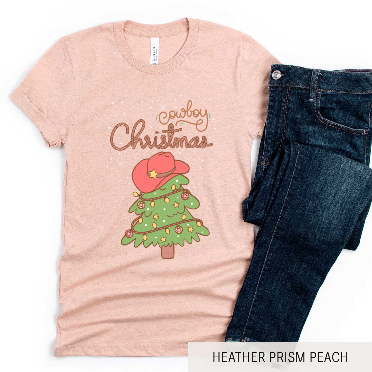 A heather prism peach Bella Canvas t-shirt featuring a cartoon christmas tree with the words cowboy christmas above it and snow falling in the background.