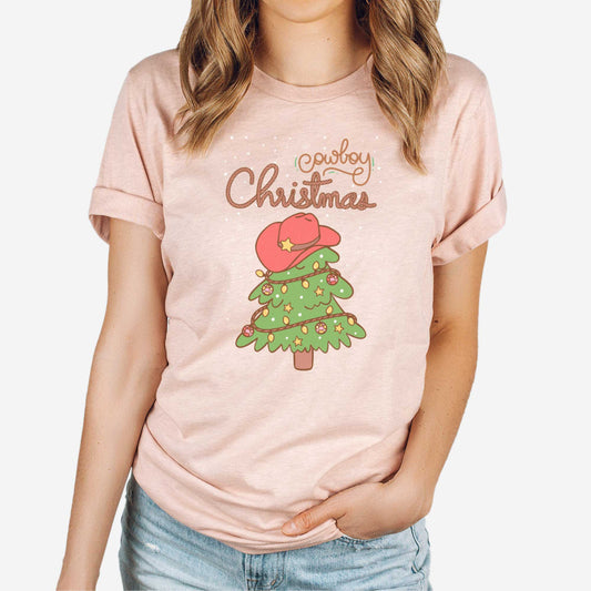 A woman wearing a heather prism peach Bella Canvas t-shirt featuring a cartoon christmas tree with the words cowboy christmas above it and snow falling in the background.