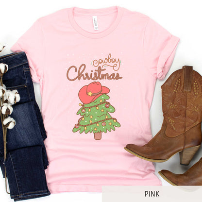 A pink Bella Canvas t-shirt featuring a cartoon christmas tree with the words cowboy christmas above it and snow falling in the background.