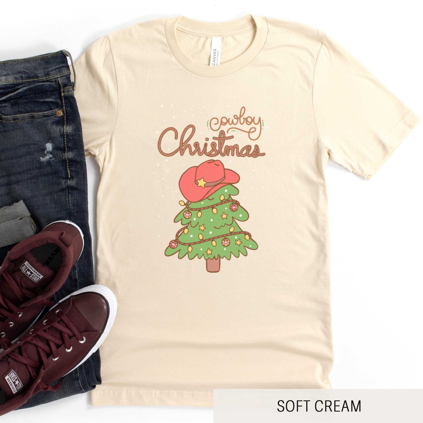 A soft cream Bella Canvas t-shirt featuring a cartoon christmas tree with the words cowboy christmas above it and snow falling in the background.