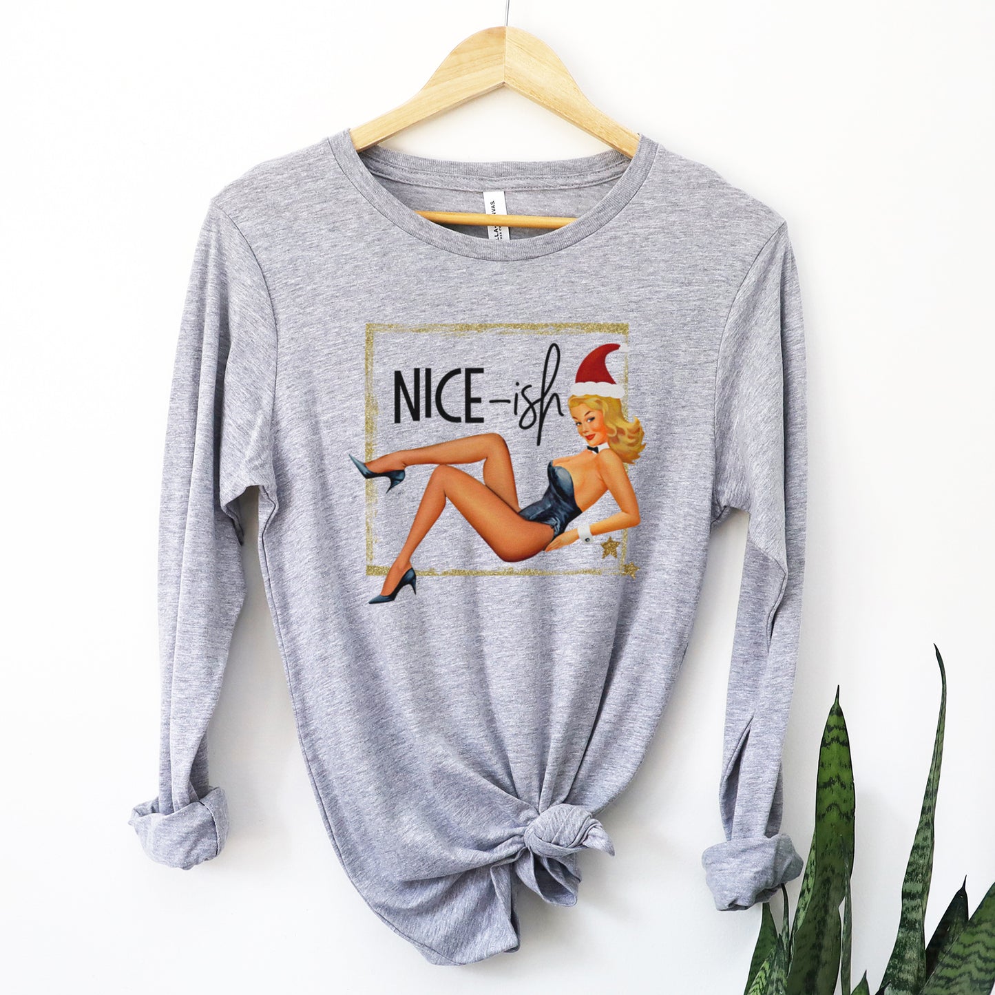 A hanging athletic heather Bella Canvas long sleeve t-shirt that features a 50's retro pin-up of a woman in a sexy outfit and stockings with a santa hat and the words nice-ish.