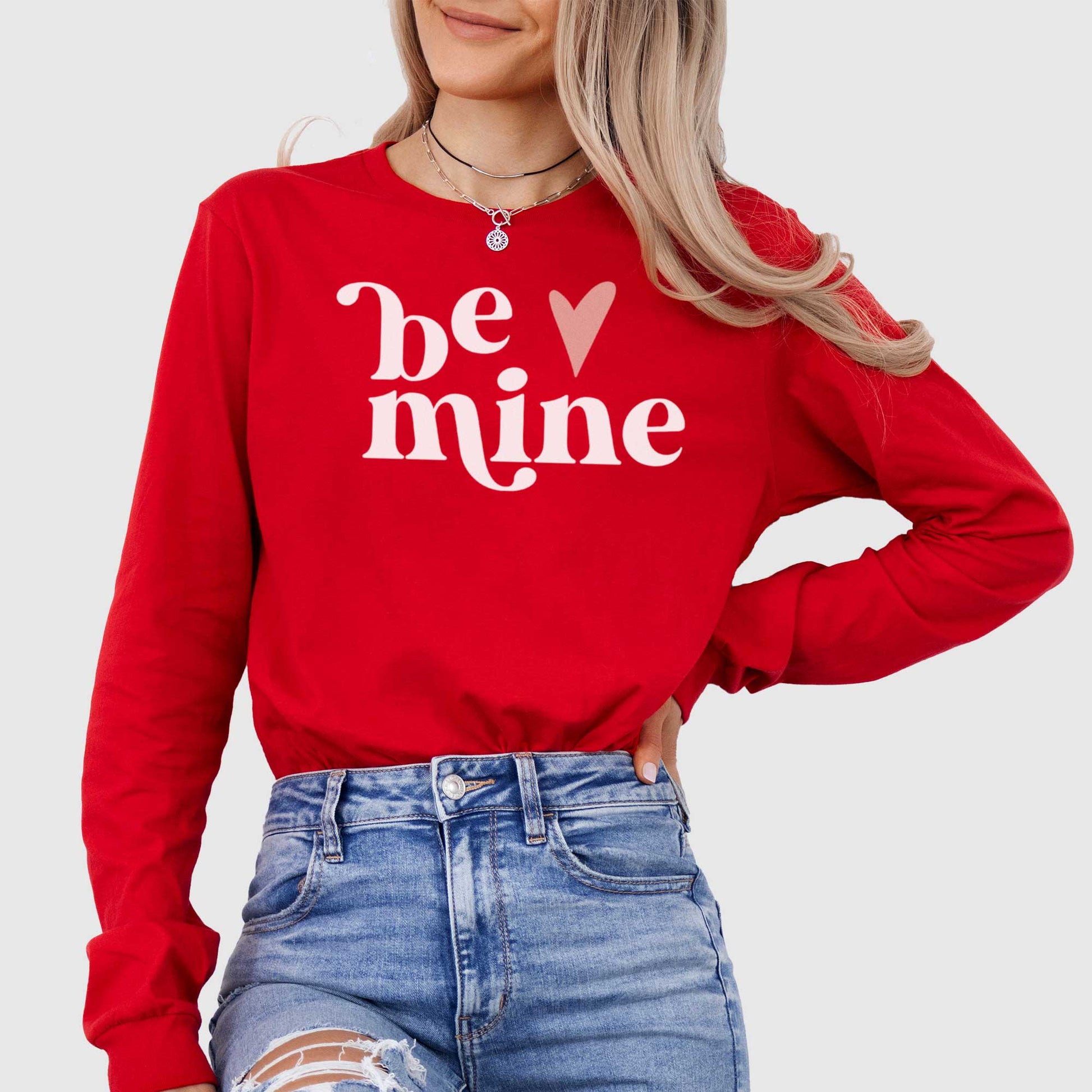 A woman wearing a red Bella Canvas long sleeve t-shirt with the words be mine and a heart next to it.