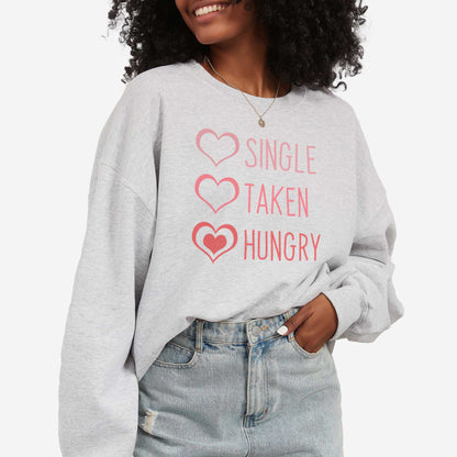 A woman wearing an ash Gildan sweatshirt featuring 3 hearts with the words single, taken, and hungry next to them.