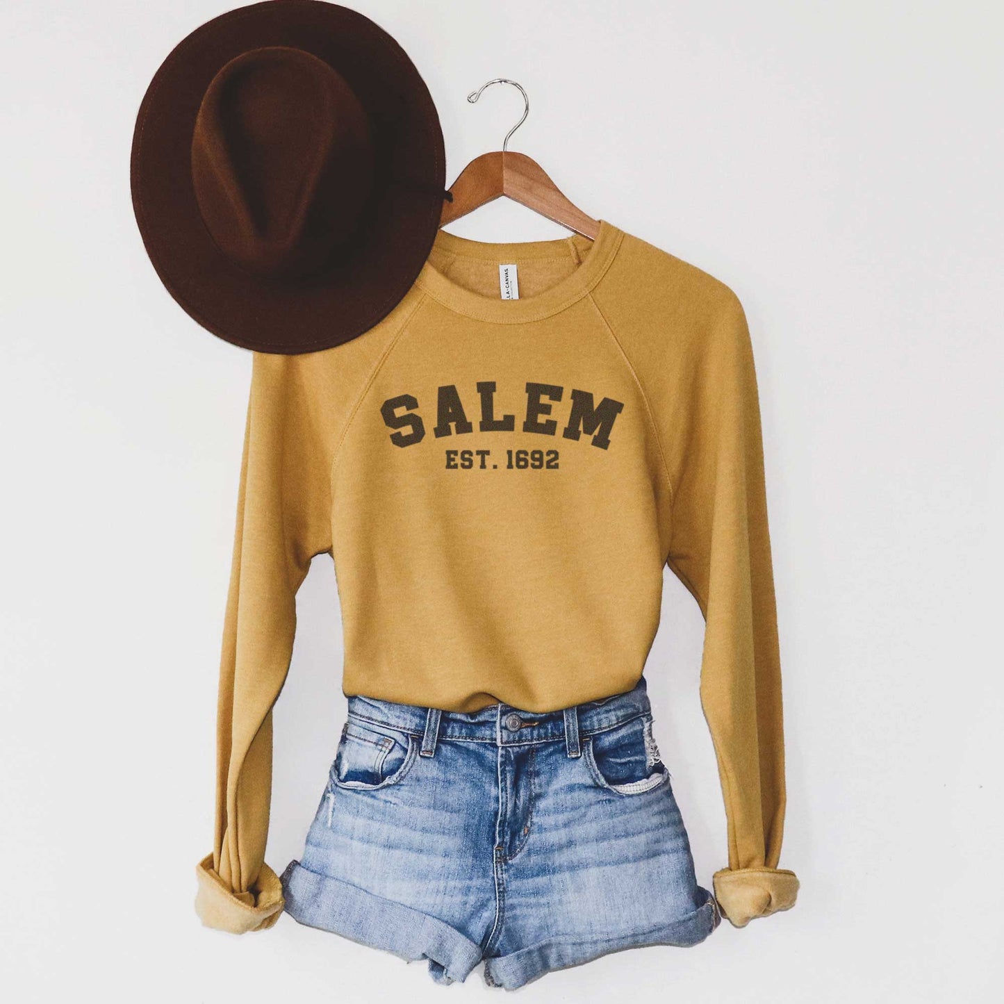 A hanging mustard yellow Bella Canvas sweatshirt featuring the words Salem Est. 1692.