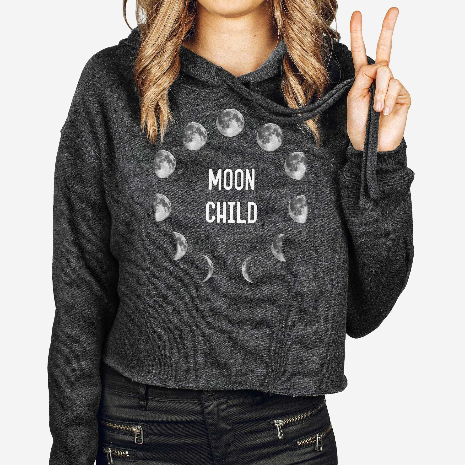 A woman wearing a cropped dark grey heather Bella Canvas hoodie featuring the phases of the moon in a circle and the words moon child.