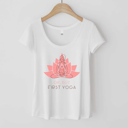 A hanging white District 7501 scoop neck t-shirt featuring a woman in the yoga lotus pose overlayed on top of a lotus flower and the words ok but first yoga.