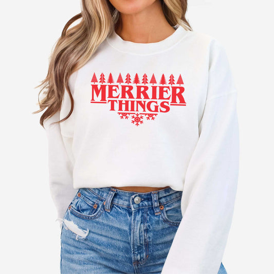 A woman wearing a white Gildan sweatshirt featuring the words merrier things surrounded by trees and snowflakes.